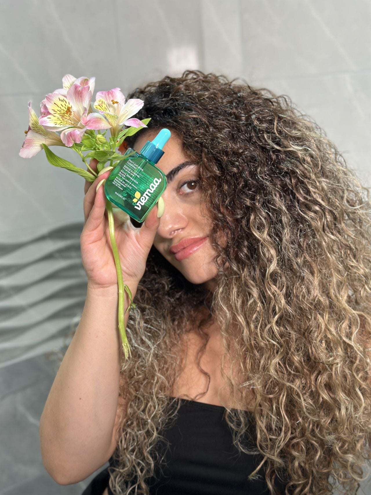 A pretty young woman holds up a flower and her frizzy hair treatment of choice: viemaa's REPAIR ELIXIR OIL. 